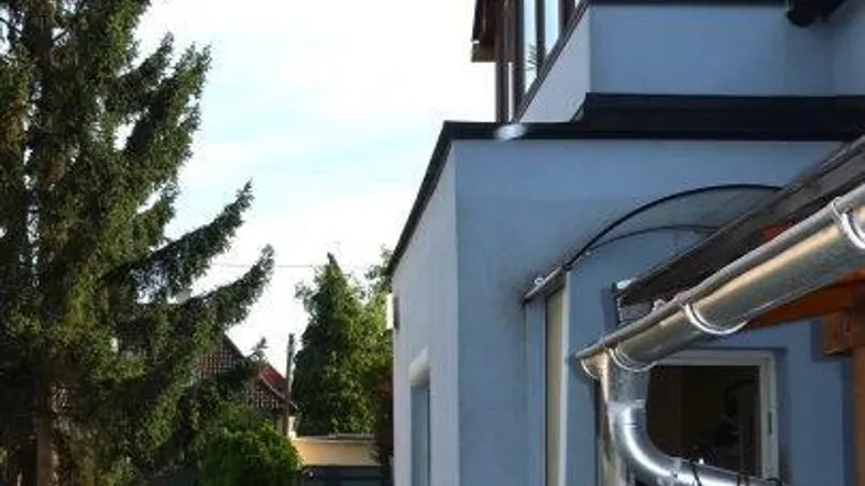 Seitenansicht eines Hauses mit hellblauer Fassade, umgeben von Bäumen und einem kleinen Gartenweg.