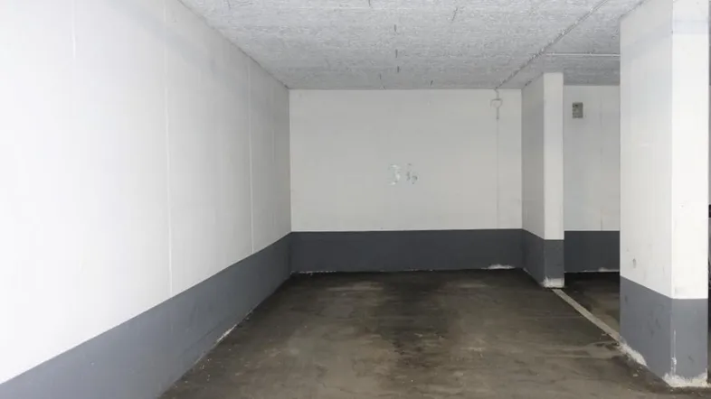 Leerer Tiefgaragenstellplatz mit weiß-grauen Wänden und sichtbaren Leitungen an der Decke.