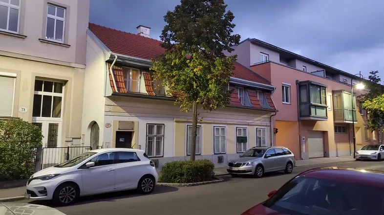 Historisches Gebäude mit Mansarddach und angrenzenden modernen Bauten in einer städtischen Umgebung.