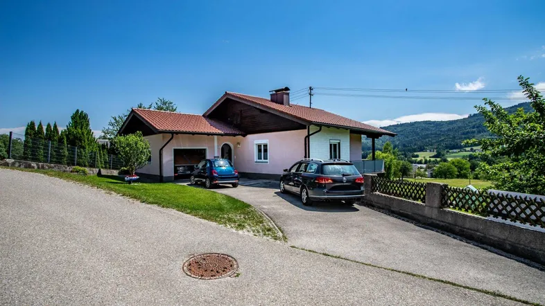 Vorderansicht des Hauses mit Einfahrt und geparkten Autos vor der grünen Hügellandschaft.