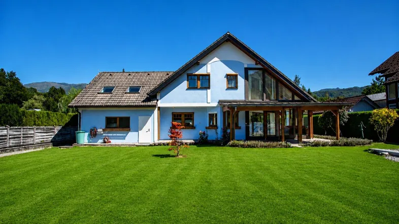 Blaues Einfamilienhaus mit großem, gepflegtem Rasen und Holzanbau unter blauem Himmel.