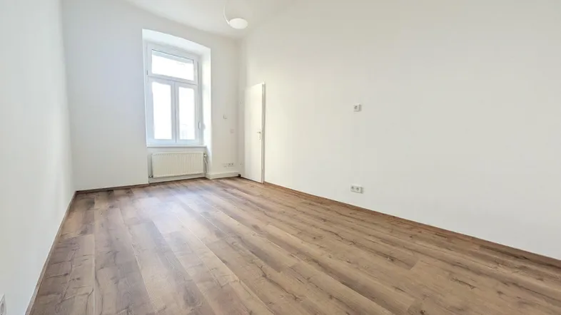 Helles, leeres Zimmer mit Laminatboden und einem Fenster, das viel Tageslicht hereinlässt.
