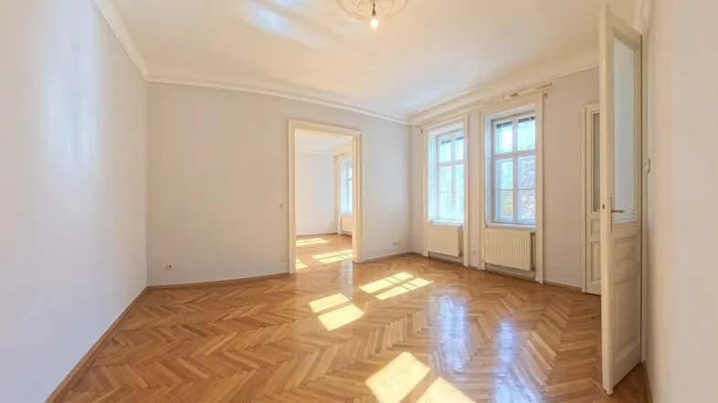 Helles Zimmer mit Fischgrätparkett, hohen Decken und großen Fenstern, das viel Tageslicht hereinlässt.