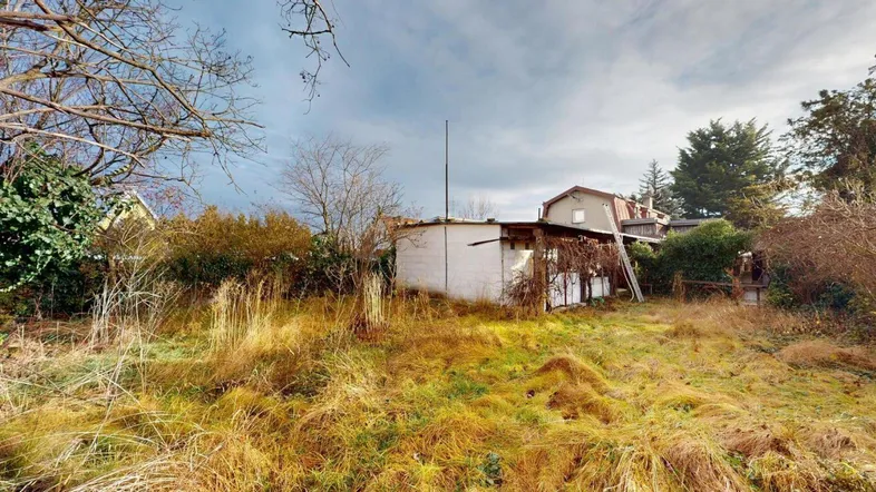 Verwilderter Gartenbereich mit einem kleinen Schuppen und Blick auf ein Wohnhaus im Hintergrund.