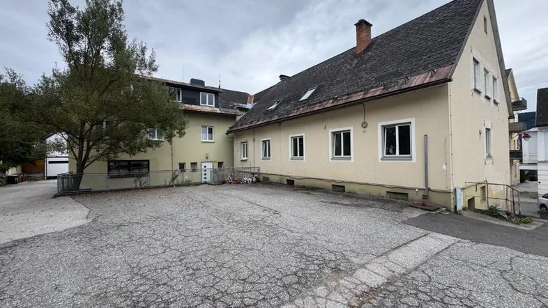 Außenansicht eines Gebäudes mit großem gepflasterten Vorplatz und einem Baum.