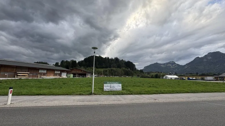 Grünes Grundstück mit einem 'Zu Verkaufen'-Schild am Straßenrand, umgeben von Bergen und bewölktem Himmel.