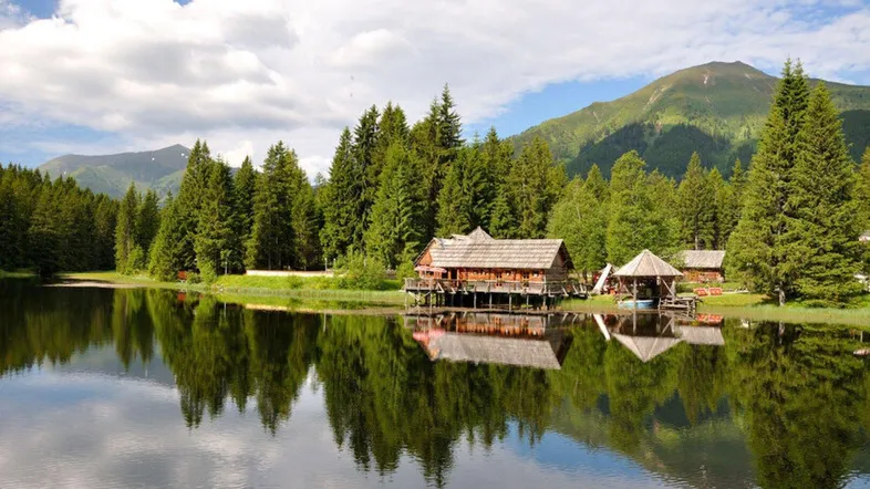 Blockhaus am Seeufer, umgeben von dichten Wäldern und Bergen, spiegelt sich im klaren Wasser.