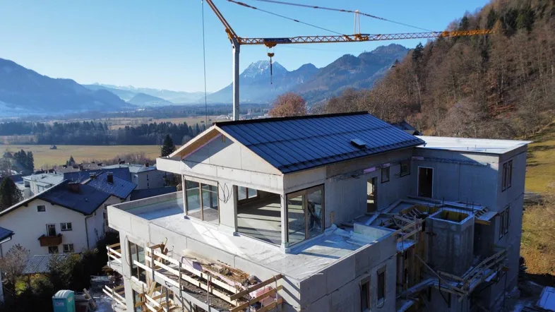 Neubau im Bau mit Kran und Bergblick, umgeben von Natur und weiteren Häusern.