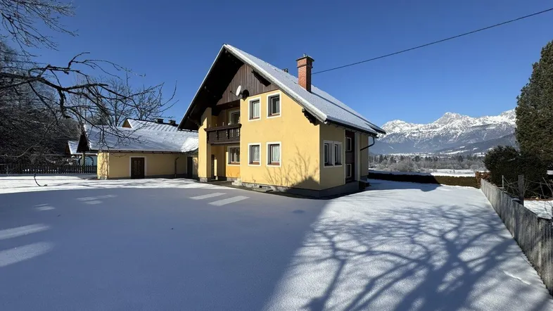 Traditionelles Haus mit gelber Fassade und schneebedecktem Dach vor einer beeindruckenden Bergkulisse.