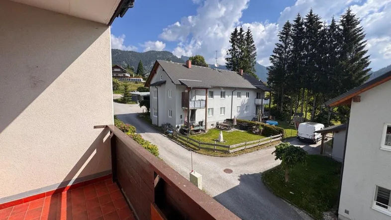 Blick vom Balkon auf eine ländliche Umgebung mit Wohngebäuden und Bergen im Hintergrund unter blauem Himmel.