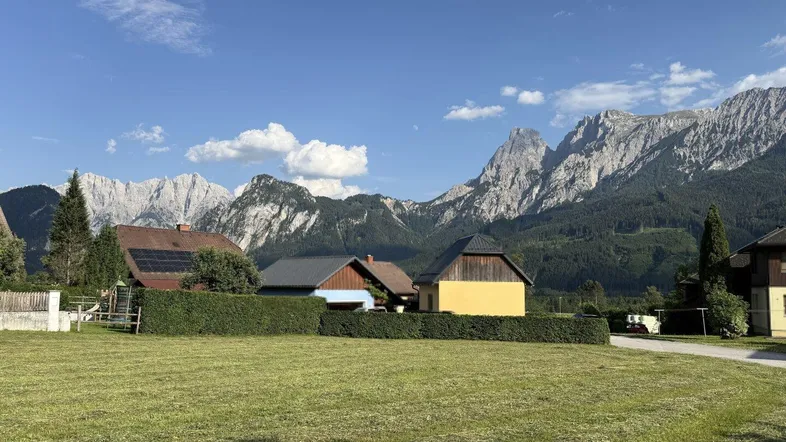 Weitläufige Wiesenfläche mit Blick auf traditionelle Häuser und majestätische Berglandschaft unter blauem Himmel.