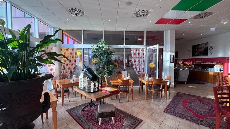 Geräumiger Restaurantbereich mit mehreren Tischen, Fliesenboden und großen Fenstern, die viel Tageslicht hereinlassen.