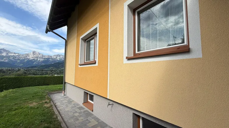 Gelbe Fassade eines Hauses mit braunen Fenstern und einem beeindruckenden Bergblick im Hintergrund.
