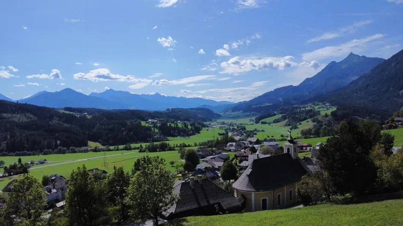 Panoramablick auf ein malerisches Dorf in einem grünen Tal, umgeben von majestätischen Bergen unter blauem Himmel.