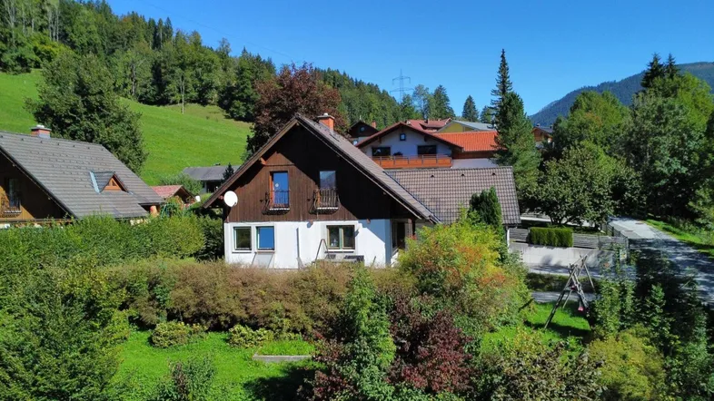 Haus in ländlicher Umgebung mit Satteldach und Holzbalkon, umgeben von viel Grün und Bergen.