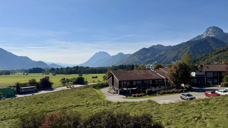 Weitläufige Landschaft mit Bergen und einem Gebäude in ländlicher Umgebung unter blauem Himmel.