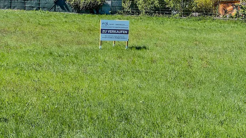 Grünes Grundstück mit einem 'Zu Verkaufen'-Schild, umgeben von Bäumen und Wohnhäusern.