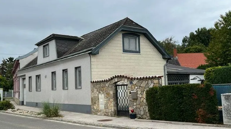Traditionelles Haus mit Satteldach und Steinfassade, umgeben von einer Hecke an der Straße.