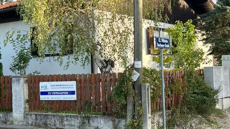 Die Außenansicht des Hauses zeigt eine weiße Fassade, einen Holzzaun und üppige Bäume, mit einem 'Zu verkaufen'-Schild im Vordergrund.