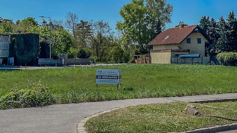 Freies Baugrundstück mit Verkaufsschild, angrenzend an einen Gehweg und umgeben von Natur.