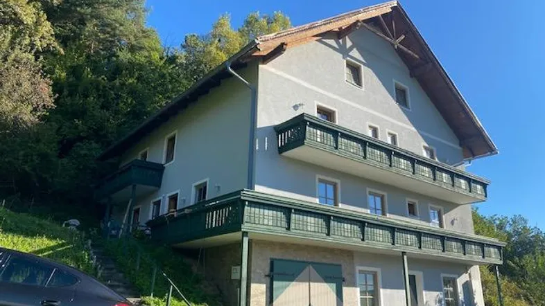 Mehrstöckiges Haus mit traditionellen Balkonen und Satteldach, eingebettet in eine grüne Hanglage.
