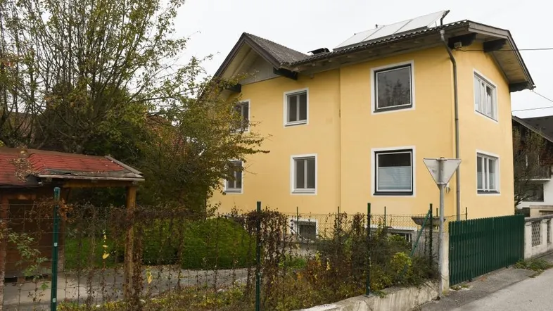 Gelbes Wohnhaus mit Satteldach und Photovoltaikanlage, umgeben von Bäumen und einem Zaun.