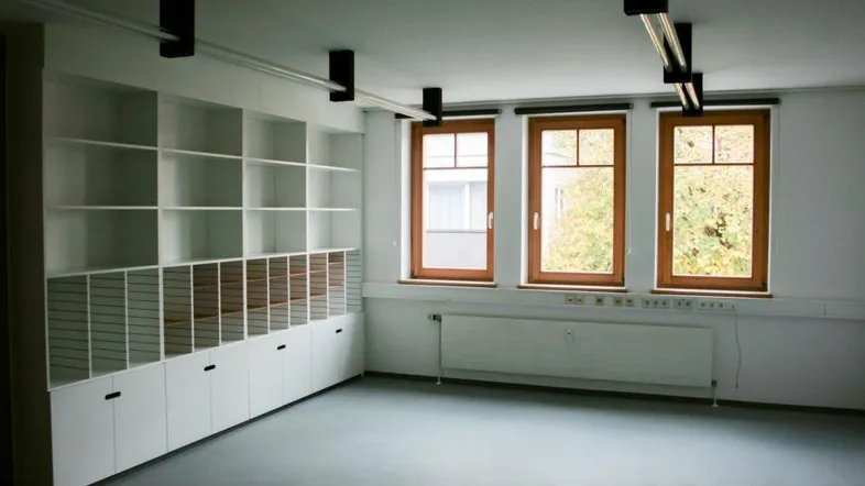 Heller Büroraum mit großen Fenstern, Einbauregalen und grauem Bodenbelag, ideal für Arbeitsbereiche.