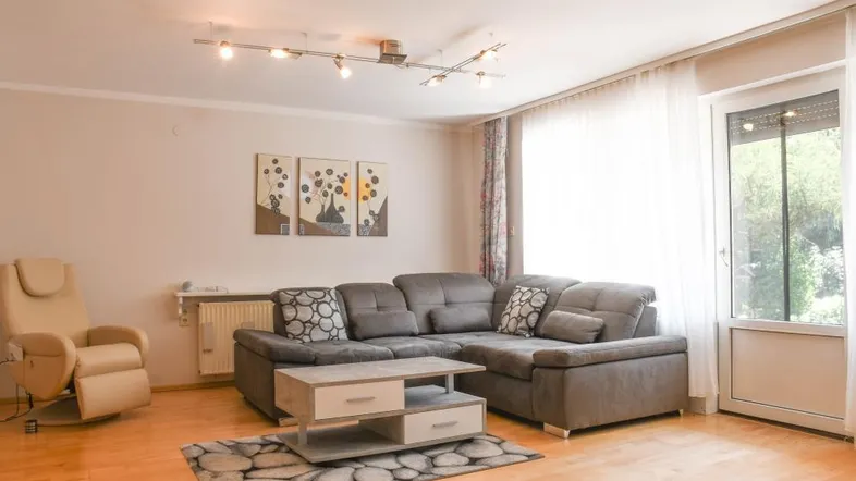 Gemütliches Wohnzimmer mit bequemer Eckcouch, Holzboden und Zugang zum Balkon.