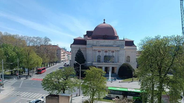Panoramablick auf ein historisches Gebäude mit Kuppel, umgeben von einer belebten Straße und grünen Bäumen unter blauem Himmel.