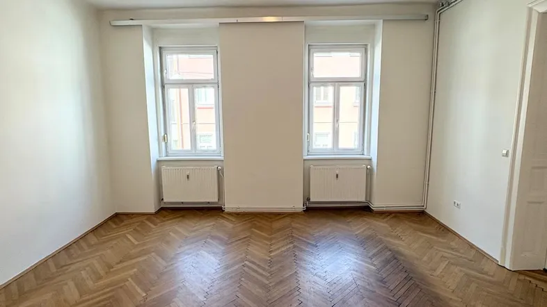 Heller Raum mit klassischem Fischgrätparkettboden und zwei Fenstern, bereit zur individuellen Gestaltung.