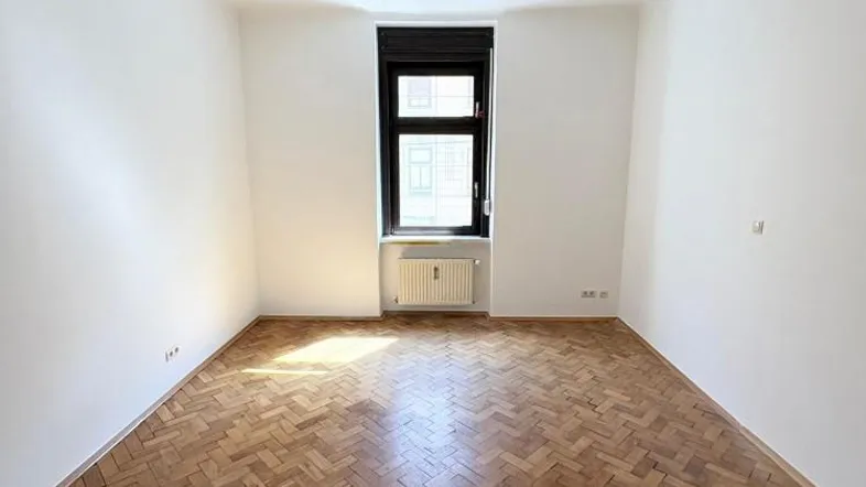 Helles Zimmer mit Fischgrätparkettboden und einem Fenster mit dunklen Rollläden.