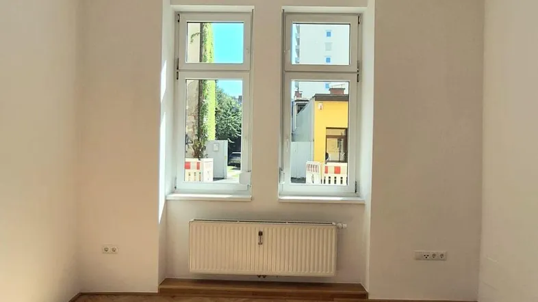 Helles Zimmer mit Fischgrätparkett und zwei Fenstern, durch die Sonnenlicht einfällt.