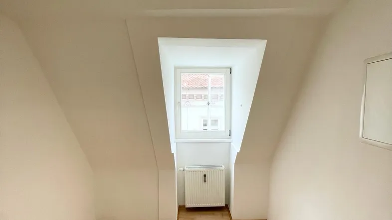 Helles Zimmer im Dachgeschoss mit schrägen Wänden, Holzboden und einem Fenster.