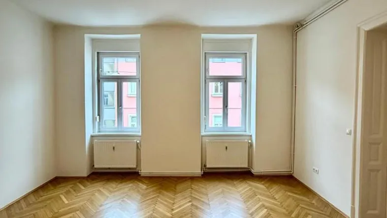 Helles, leeres Zimmer mit Fischgrätparkettboden und zwei Fenstern, ideal als Wohnzimmer nutzbar.