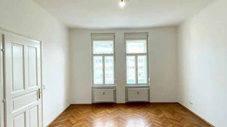Heller Raum mit Fischgrätparkettboden, weißen Wänden und zwei hohen Fenstern.
