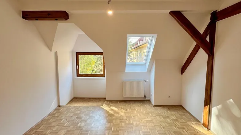 Heller Dachgeschossraum mit Parkettboden, sichtbaren Holzbalken und schrägen Decken.
