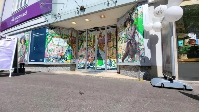 Eingangsbereich eines Geschäftsgebäudes mit großen Glasflächen und Werbeplakaten, die die Fassade bedecken.