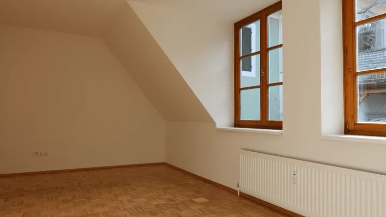 Helles Zimmer im Dachgeschoss mit Parkettboden, schrägen Wänden und zwei Holzfenstern.