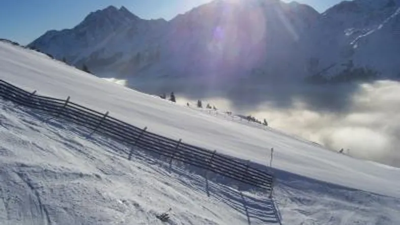 Sonniger Skihang mit verschneiten Bergen und Wolkenmeer im Tal, ideal für Wintersport.