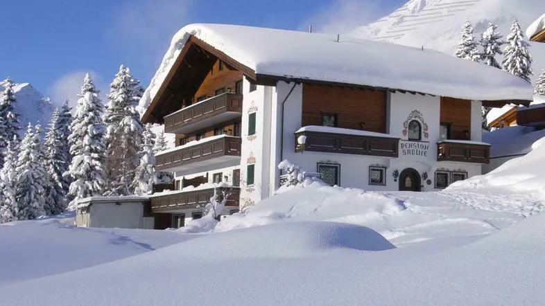 Traditionelles Gebäude im alpinen Stil, komplett mit Schnee bedeckt, umgeben von verschneiten Bergen und Tannen.