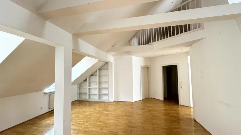 Geräumiger Wohnbereich im Dachgeschoss mit Holzboden, sichtbaren Balken und integriertem Regal unter der Dachschräge.
