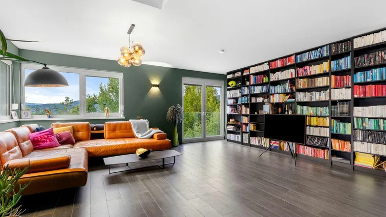 Geräumiges Wohnzimmer mit Ledersofa, großer Bücherwand, dunklem Holzboden und Blick ins Grüne.
