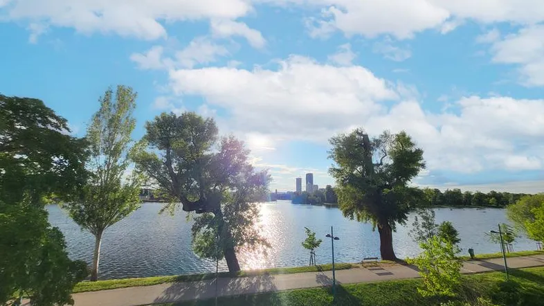 Panoramablick auf einen See mit Bäumen und einer Stadtkulisse in der Ferne unter blauem Himmel.
