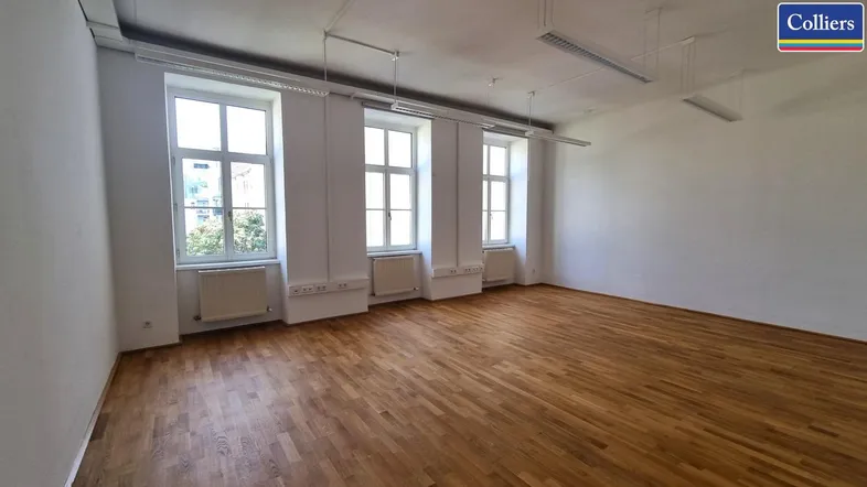Geräumiger, leerer Büroraum mit Holzboden und großen Fenstern, die viel Tageslicht hereinlassen.