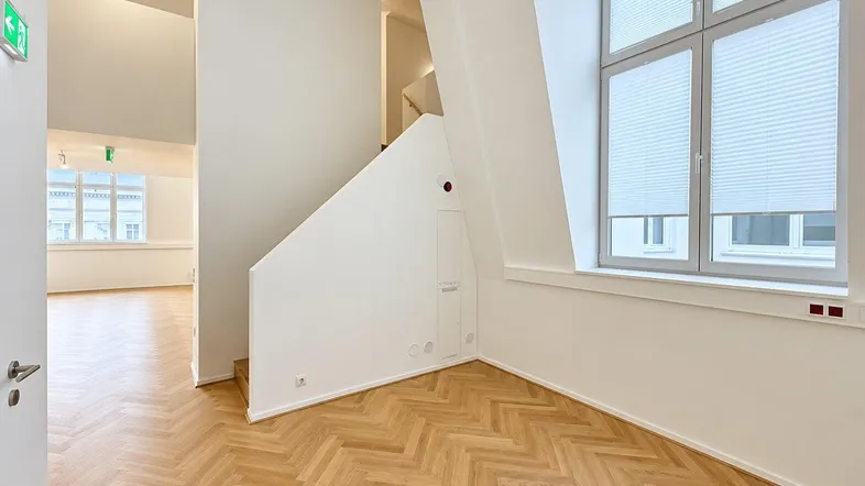 Heller Innenraum mit Fischgrätparkett, weiß gestrichenen Wänden und einer modernen Treppe, die in den oberen Bereich führt.