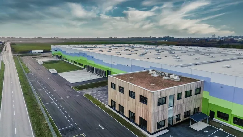 Luftaufnahme eines modernen Logistikzentrums mit angrenzendem Bürogebäude und weitläufigen Parkplätzen unter bewölktem Himmel.