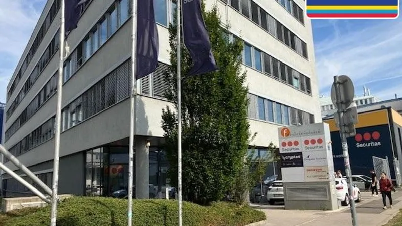 Modernes Bürogebäude mit mehreren Etagen und umliegender Grünfläche, sichtbar unter blauem Himmel.