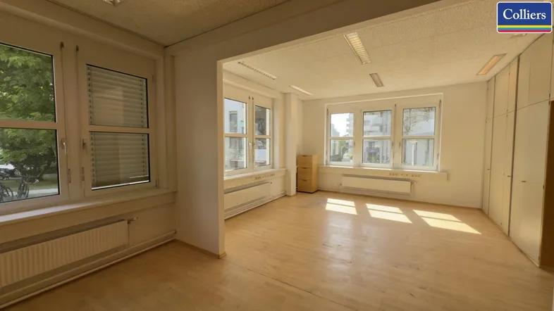 Geräumiges, leeres Büro mit hellem Holzfußboden und großen Fenstern, die viel Tageslicht hereinlassen.