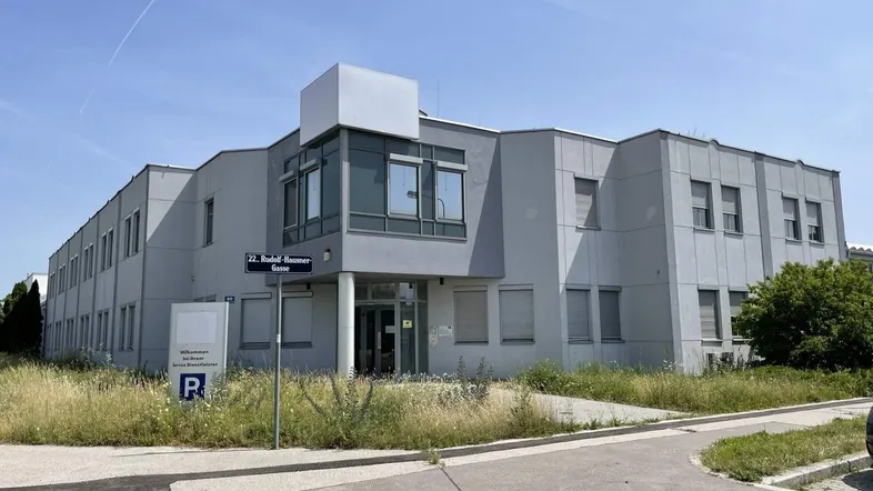 Modernes Bürogebäude mit grauer Fassade und Parkplatz im Vordergrund unter blauem Himmel.