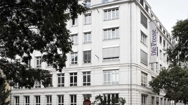 Ein klassisches, weißes Bürogebäude mit vielen Fenstern und Jalousien, umgeben von Bäumen in einer städtischen Umgebung.